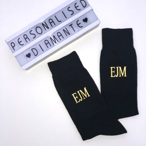 May include: A pair of black socks with gold embroidered initials "EJM". The socks are displayed on a white background with a white light-up sign that reads "PERSONALISED &hearts; DIAMANTE".