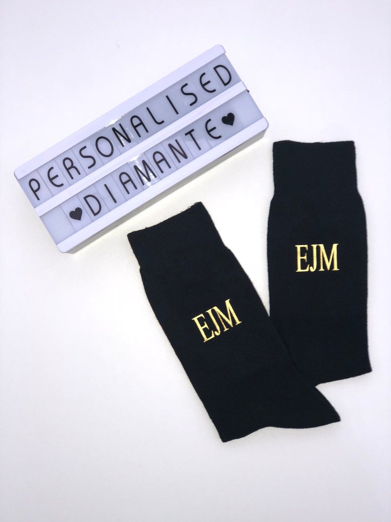 May include: A pair of black socks with gold embroidered initials "EJM". The socks are displayed on a white background with a white light-up sign that reads "PERSONALISED &hearts; DIAMANTE".