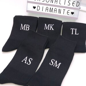 May include: Five pairs of black socks with white embroidered initials: MB, MK, TL, AS, and SM. The socks are arranged in a semi-circle on a white surface. The text "Personalised Diamante" is visible in the background.