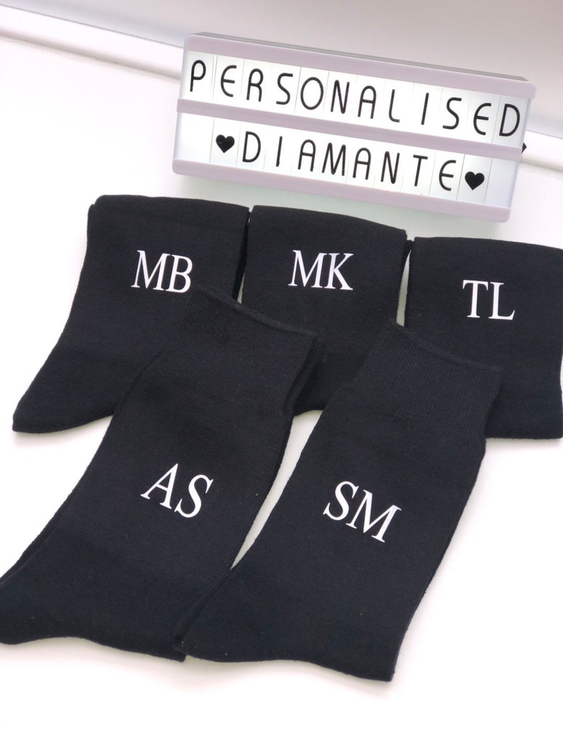 May include: Five pairs of black socks with white embroidered initials: MB, MK, TL, AS, and SM. The socks are arranged in a semi-circle on a white surface. The text "Personalised Diamante" is visible in the background.