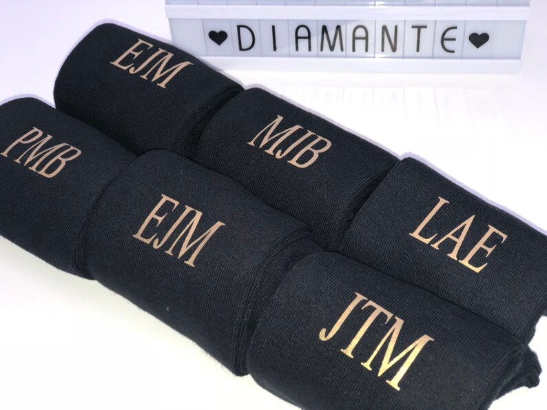 May include: Six black fabric wrist cuffs with gold embroidered initials. The initials are EJM, PMB, MJB, LAE, and JTM.