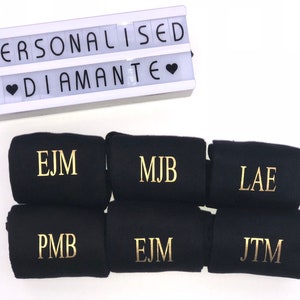 May include: Six black socks with gold embroidered initials. The initials are EJM, MJB, LAE, PMB, EJM, and JTM. The socks are arranged in two rows of three. A white light box with the words "Personalised Diamante" is in the background.