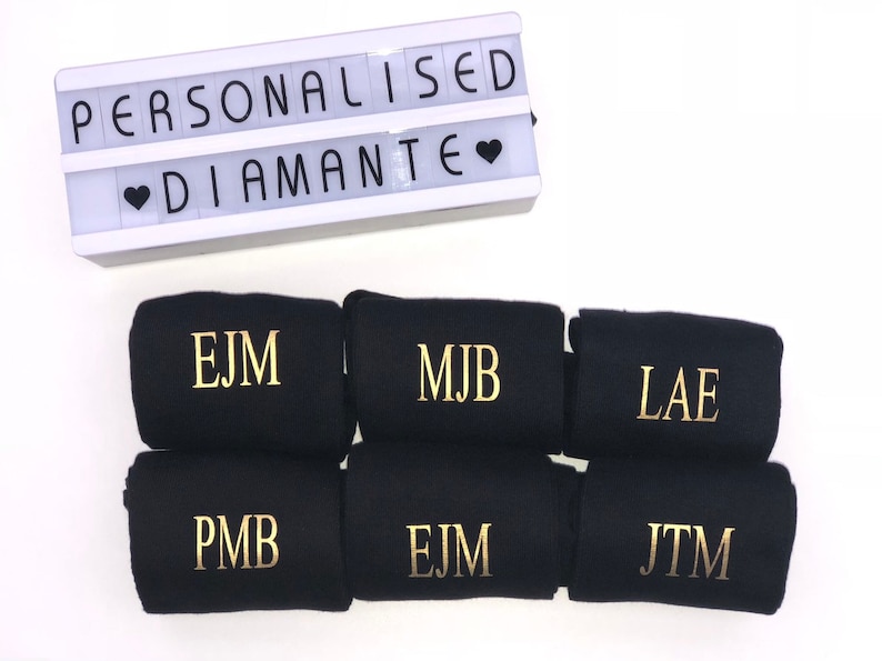 May include: Six black socks with gold embroidered initials. The initials are EJM, MJB, LAE, PMB, EJM, and JTM. The socks are arranged in two rows of three. A white light box with the words "Personalised Diamante" is in the background.