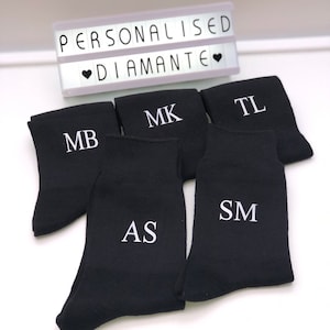 May include: Five pairs of black socks with white embroidered initials. The initials are MB, MK, TL, AS, and SM. The socks are arranged in a semi-circle on a white surface. A light box with the text "PERSONALISED &hearts; DIAMANTE &hearts;" is visible in the background.