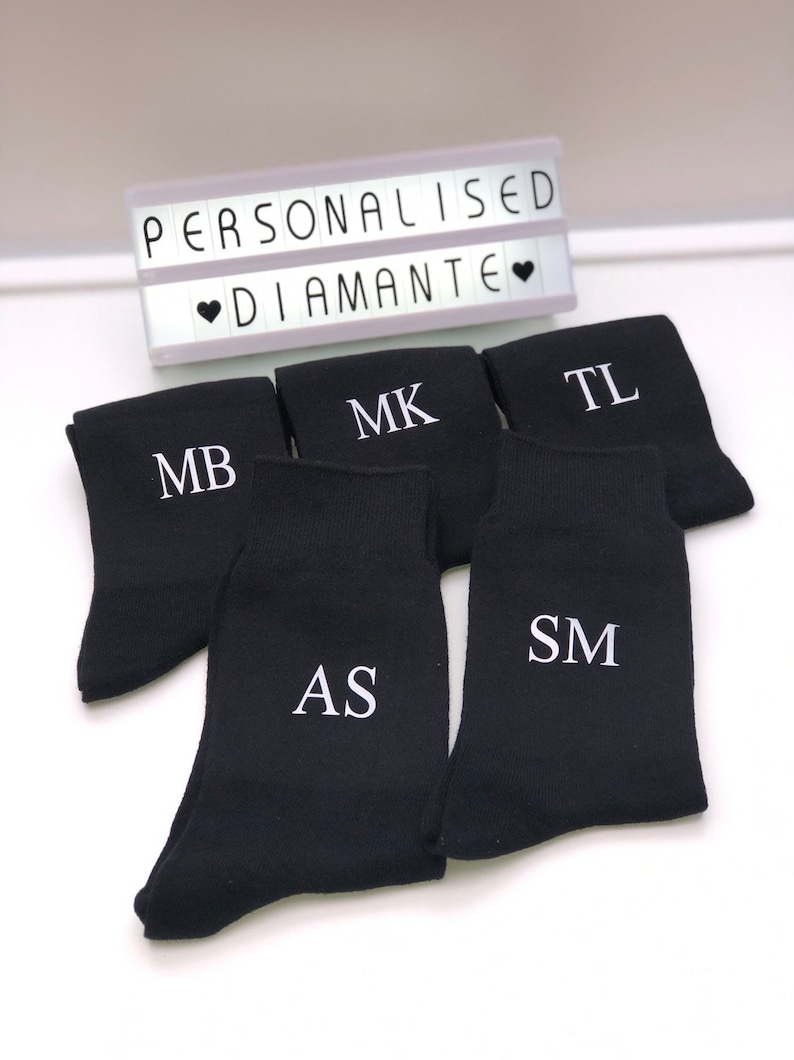 May include: Five pairs of black socks with white embroidered initials. The initials are MB, MK, TL, AS, and SM. The socks are arranged in a semi-circle on a white surface. A light box with the text "PERSONALISED &hearts; DIAMANTE &hearts;" is visible in the background.