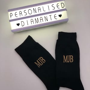 May include: A pair of black socks with the initials "MJB" embroidered in gold on each sock. A light box with the words "Personalised Diamante" is in the background.