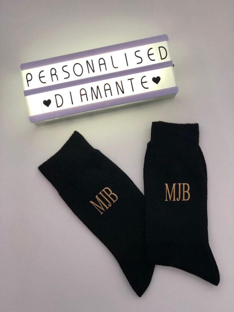 May include: A pair of black socks with the initials "MJB" embroidered in gold on each sock. A light box with the words "Personalised Diamante" is in the background.
