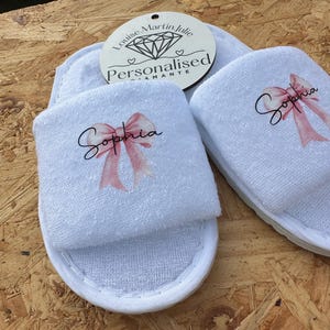 Pode incluir: Um par de chinelos brancos de tecido felpudo com uma fita rosa e o nome "Sophia" bordado em cada chinelo. Os chinelos estão sobre uma superfície de madeira.