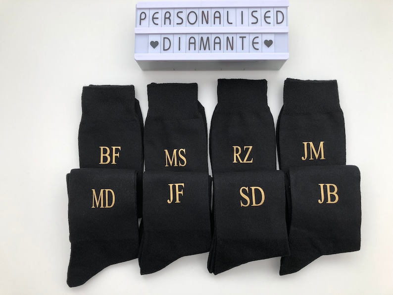May include: Eight pairs of black socks with gold embroidered initials. The socks are arranged in two rows of four. The socks are displayed on a white surface. The text "PERSONALISED DIAMANTE" is visible in the background.