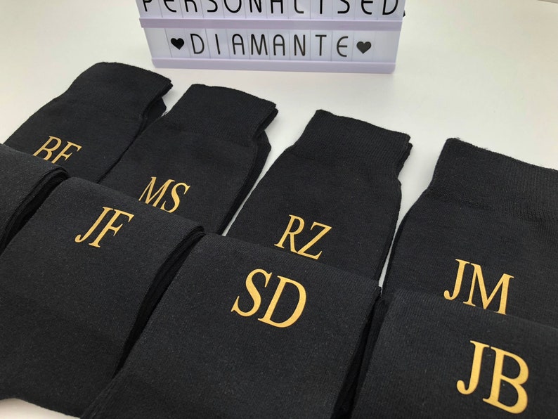 May include: A set of eight black socks with gold embroidered initials. The initials are: RF, JF, MS, RZ, SD, JM, and JB.