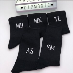 May include: Five pairs of black socks with white embroidered initials. The initials are MB, MK, TL, AS, and SM.