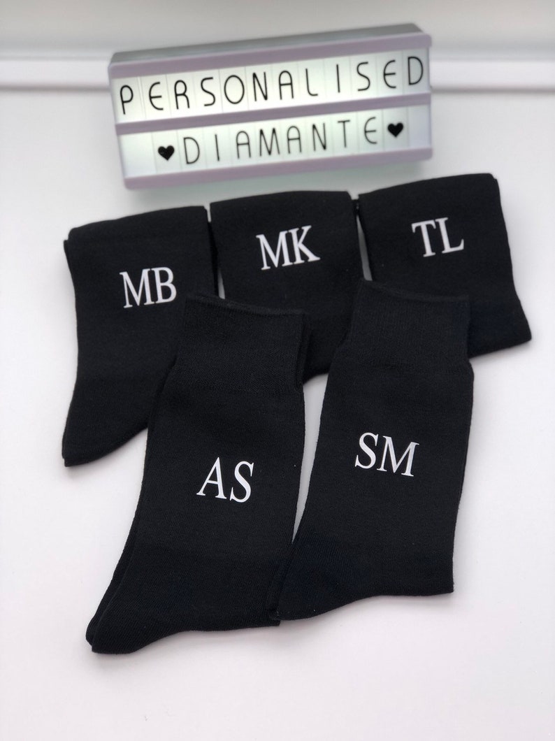 May include: Five pairs of black socks with white embroidered initials. The initials are MB, MK, TL, AS, and SM.