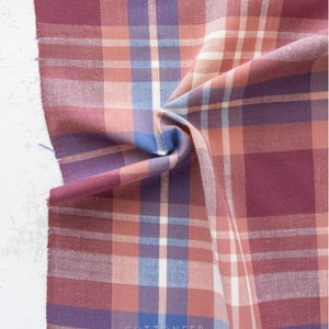 May include: Close-up of a fabric with a plaid pattern. The fabric features a mix of colors, including burgundy, light blue, peach, and white. The word "COTTONEER" is printed on the fabric. The fabric is folded and has a raw edge.