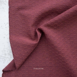May include: Close-up of a burgundy fabric with a textured, geometric pattern. The fabric is folded, showing depth and dimension. The word "FABLEISM" is printed in white on the fabric.