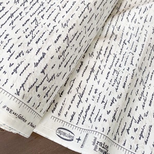 May include: A roll of fabric with a cream-colored background and a handwritten script pattern in dark gray. The fabric is labeled with text including "#WE ARE fabrics" and "capsules collection". The fabric is from "AGF Studio".