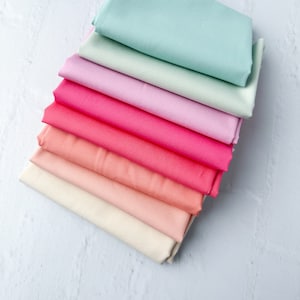 May include: A stack of eight fabric squares in various shades of pink, peach, and light blue. The fabric is folded in half and arranged in a gradient from light blue to pink.
