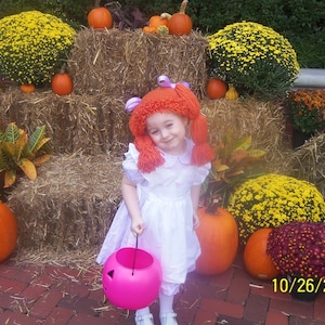 May include: A young girl dressed as a character from the movie "The Wizard of Oz" with a red wig and a white dress. She is holding a pink pumpkin bucket and is standing in front of a hay bale backdrop with pumpkins and fall foliage.