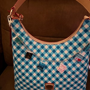 May include: A blue and white gingham patterned handbag with a pink strap and gold hardware. A silver chain with book charms hangs from the zipper. The charms have book titles like "Ugly Love" and "November 9".