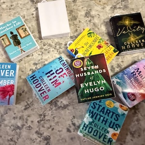 May include: A collection of miniature books with various covers, including titles like "Verity" and "November 9". The books are in different colors, with visible text and author names. A white gift bag is also present.