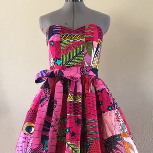May include: A strapless, patchwork dress in shades of pink, with a fitted bodice and a flared skirt. The dress is decorated with various colourful patterns, including stars, leaves, and geometric shapes. A matching fabric belt cinches the waist, creating a flattering silhouette.