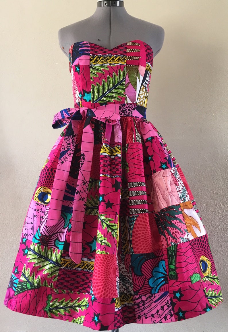 May include: A strapless, patchwork dress in shades of pink, with a fitted bodice and a flared skirt. The dress is decorated with various colourful patterns, including stars, leaves, and geometric shapes. A matching fabric belt cinches the waist, creating a flattering silhouette.