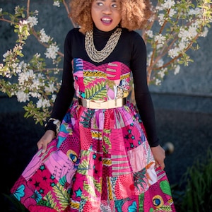 May include: A colourful patchwork dress with a strapless bodice and a full skirt. The dress features a mix of pink, purple, and green patterned fabrics, cinched at the waist with a gold-tone belt. A black long-sleeved top is worn underneath.
