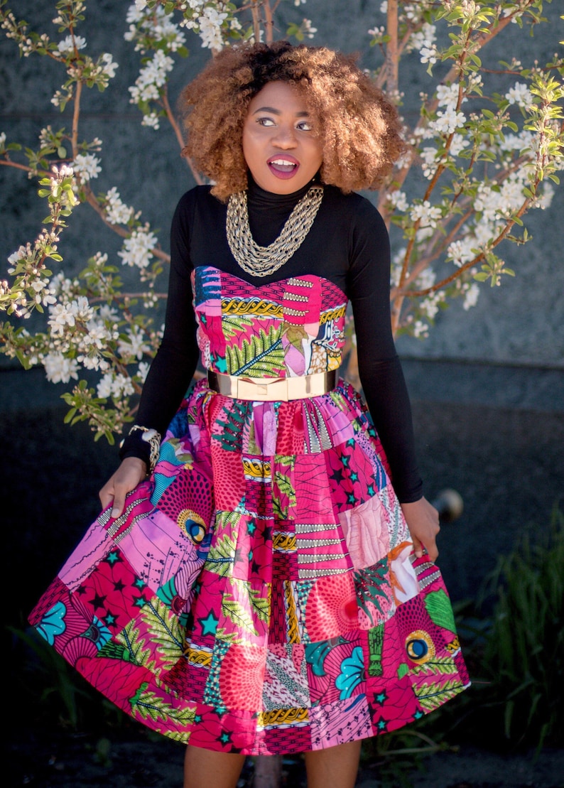 May include: A colourful patchwork dress with a strapless bodice and a full skirt. The dress features a mix of pink, purple, and green patterned fabrics, cinched at the waist with a gold-tone belt. A black long-sleeved top is worn underneath.