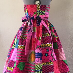 May include: A strapless patchwork dress in bright pink, green, blue, and yellow. The dress has a fitted bodice and a full skirt, with a matching fabric belt at the waist. The fabric is patterned with geometric shapes, stars, and floral designs.