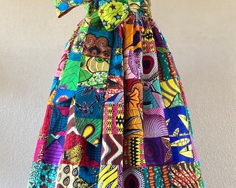 May include: A vibrant, patchwork-style maxi dress on a mannequin. The dress features a one-shoulder design with a bow at the waist. The fabric is a mix of colorful African prints in various patterns and hues, including red, green, blue, and purple.
