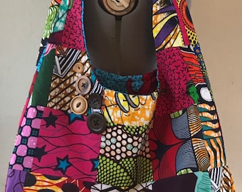 Puede incluir: Un bolso de hombro patchwork colorido con una variedad de telas estampadas en tonos rosa, morado, verde, amarillo y azul. El bolso tiene una sola correa y está adornado con varios botones de madera.