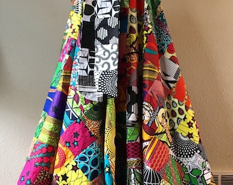 May include: A long, colorful patchwork skirt with a black and white patterned sash. The skirt is made of various African print fabrics in shades of red, yellow, green, blue, and purple.