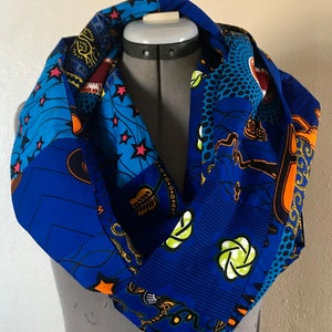 Puede incluir: Una bufanda infinita azul con un estampado africano colorido. El estampado incluye estrellas, hojas y formas geométricas en tonos naranja, amarillo, verde y negro.