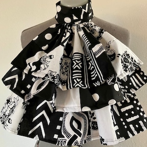 Op de afbeelding: Een zwart-wit gepatroneerde kraag van stof met meerdere lagen en een gerimpeld ontwerp. De stof heeft geometrische vormen, abstracte ontwerpen en bloemmotieven.