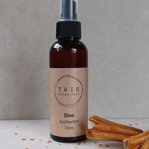 Puede incluir: Una botella de vidrio marr&oacute;n con repelente de insectos "Shoo" de Twig Botanicals, con un pulverizador negro y una etiqueta beige. La etiqueta incluye el texto "Bug Repellent" y "125ml". Vainas de semillas secas en primer plano.
