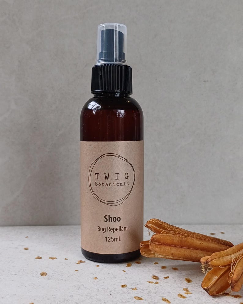 Puede incluir: Una botella de vidrio marr&oacute;n con repelente de insectos "Shoo" de Twig Botanicals, con un pulverizador negro y una etiqueta beige. La etiqueta incluye el texto "Bug Repellent" y "125ml". Vainas de semillas secas en primer plano.