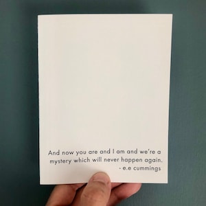 May include: A white card with black text that reads, "And now you are and I am and we're a mystery which will never happen again. - e.e cummings." The card is held against a dark teal background.