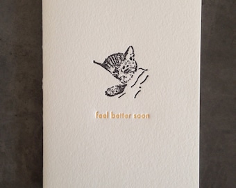 May include: A white card with a black and white illustration of a sleeping kitten. The text "feel better soon" is printed in gold on the card.