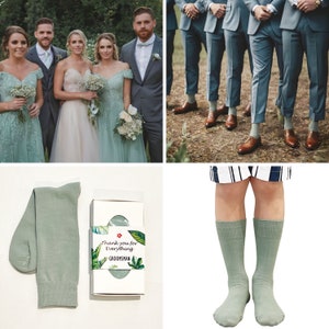 May include: A pair of sage green crew socks next to a white gift box with a tropical leaf print label that reads Thank you for Everything GROOMSMAN. The socks are also shown worn on lower legs with white and navy blue striped shorts.