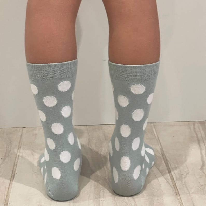 May include: A pair of mid-calf socks in a soft sage green colour featuring a repeating pattern of large, circular white polka dots. The socks have a ribbed cuff at the top and are designed to cover the lower leg, extending down to the heel area.