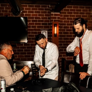 May include: Three men in a dimly lit room with a brick wall background. Two men wear white dress shirts and ties, one with a black tie and the other with a red tie. The third man wears a tan blazer.