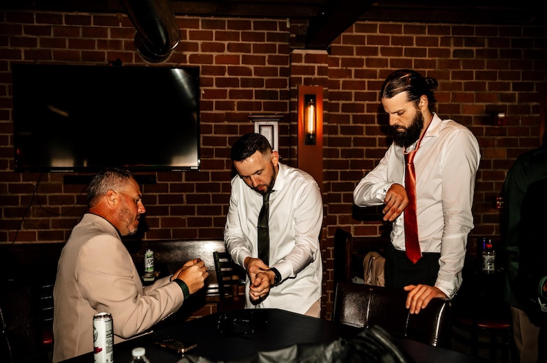 May include: Three men in a dimly lit room with a brick wall background. Two men wear white dress shirts and ties, one with a black tie and the other with a red tie. The third man wears a tan blazer.