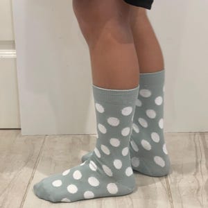 May include: A pair of mid-calf socks in a muted sage green colour featuring a repeating pattern of large, solid white polka dots. The socks have a ribbed cuff at the top and cover the feet and lower legs, showcasing a consistent dot design throughout.