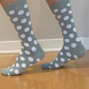 May include: A pair of crew-length socks in a muted sage green colour featuring a repeating pattern of solid white polka dots. The socks have a ribbed cuff at the top and cover the lower leg, extending down to the heel and toe area of the feet.