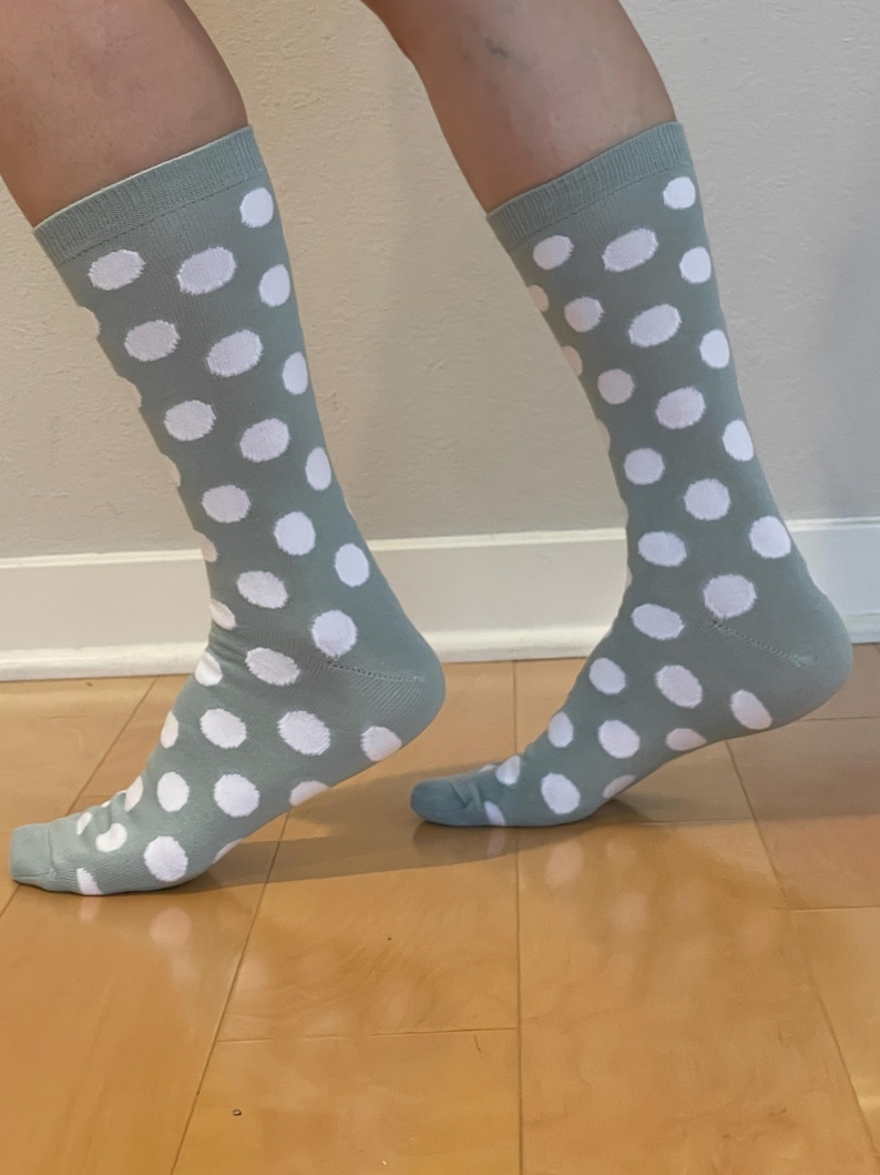 May include: A pair of crew-length socks in a muted sage green colour featuring a repeating pattern of solid white polka dots. The socks have a ribbed cuff at the top and cover the lower leg, extending down to the heel and toe area of the feet.
