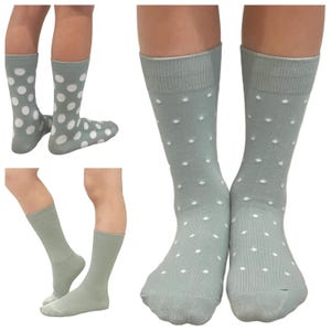 May include: A pair of mid-calf socks in a muted sage green colour featuring a repeating pattern of small white polka dots. The socks have a ribbed cuff at the top and are shown from multiple angles, including a front view and a side profile view.