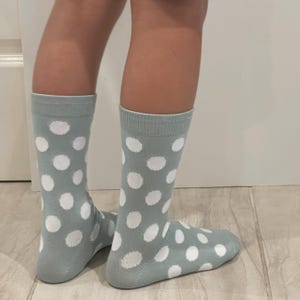 May include: A pair of mid-calf socks in a muted sage green colour featuring a repeating pattern of solid white polka dots. The socks have a ribbed cuff at the top and cover the lower leg and foot, displaying a consistent dot design throughout.