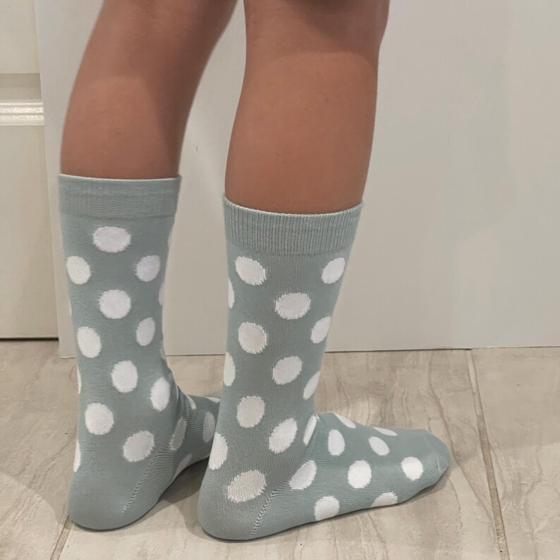 May include: A pair of mid-calf socks in a muted sage green colour featuring a repeating pattern of solid white polka dots. The socks have a ribbed cuff at the top and cover the lower leg and foot, displaying a consistent dot design throughout.