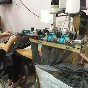 May include: An industrial sock knitting machine in operation. The machine is silver and blue, with multiple spools of thread and a small monitor. A person is holding a gray sock, and a pile of gray socks is visible.