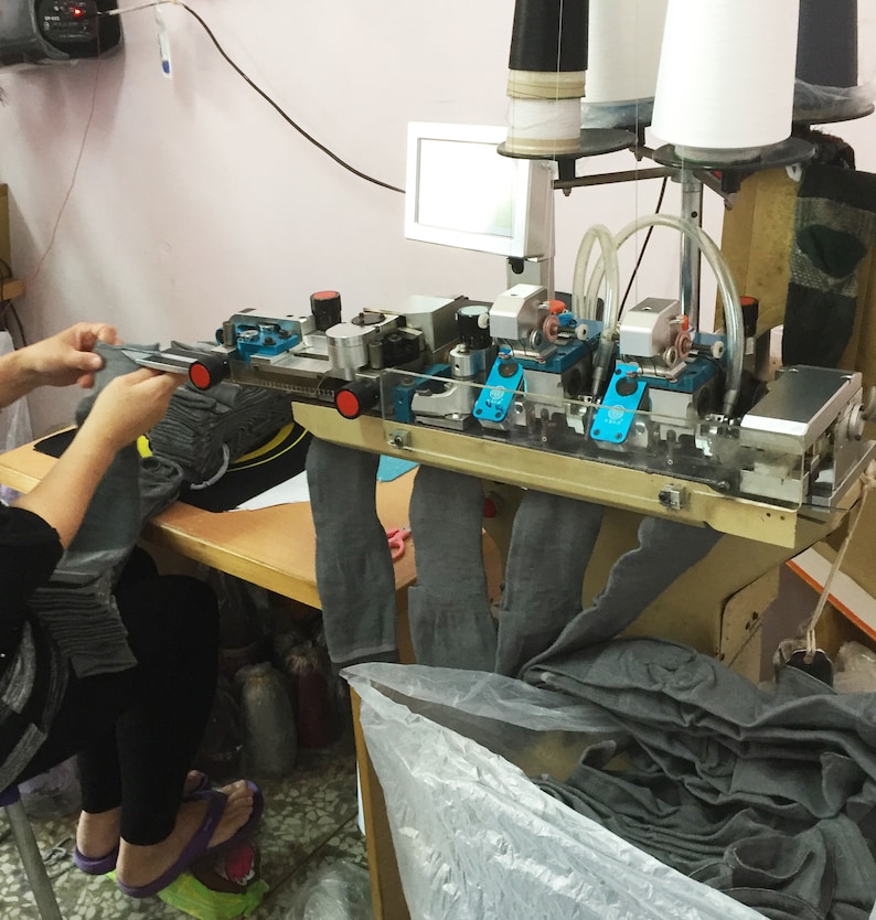 May include: An industrial sock knitting machine in operation. The machine is silver and blue, with multiple spools of thread and a small monitor. A person is holding a gray sock, and a pile of gray socks is visible.