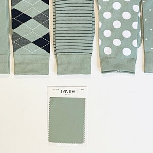 May include: Five pairs of sage green socks featuring various patterns including solid, argyle with navy and grey, horizontal stripes, large white polka dots, and small white dots, alongside a David's Bridal fabric swatch card in sage green.